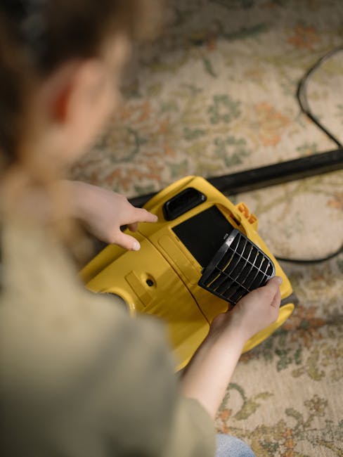 A person using a yellow portable carpet cleaning machine on a patterned rug in a room with natural lighting. The machine includes a handle, a vented cover, and a control panel, and is positioned on a 
