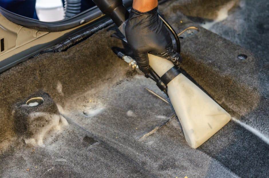 A professional cleaner dressed in a full white protective suit, including a hood, mask, and orange gloves, is operating a vacuum cleaner for surface cleaning and hygiene maintenance in a modern indoor